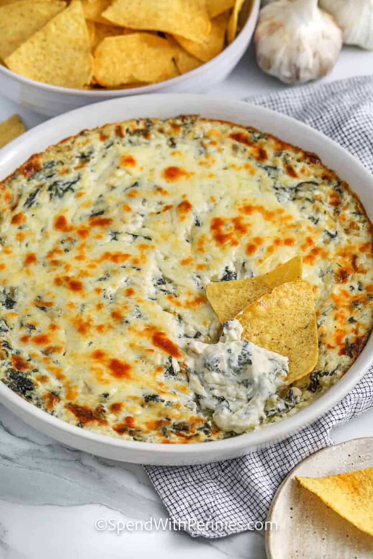 Spinach artichoke dip is creamy, cheesy, and full of flavor in every bite. It’s made with cream cheese, sour cream, and a mix of melted cheeses, then baked until hot and golden. This easy recipe pairs perfectly with bread, chips, or vegetables. It’s one of the best appetizers to make for parties or family dinners. #spendwithpennies baked spinach artichoke dip with two tortilla chips on top