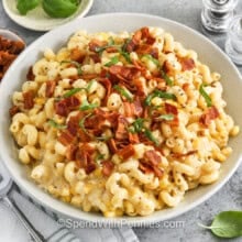 serving bowl of creamy corn pasta with a serving spoon on the side, and small bowls of bacon, basil, and red pepper flakes