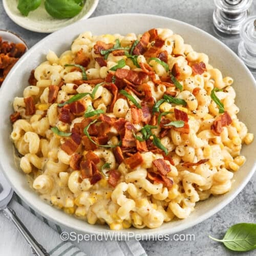 serving bowl of creamy corn pasta with a serving spoon on the side, and small bowls of bacon, basil, and red pepper flakes