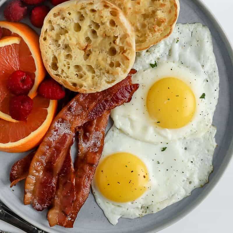 fried eggs on plate with english muffin, bacon, and fruit