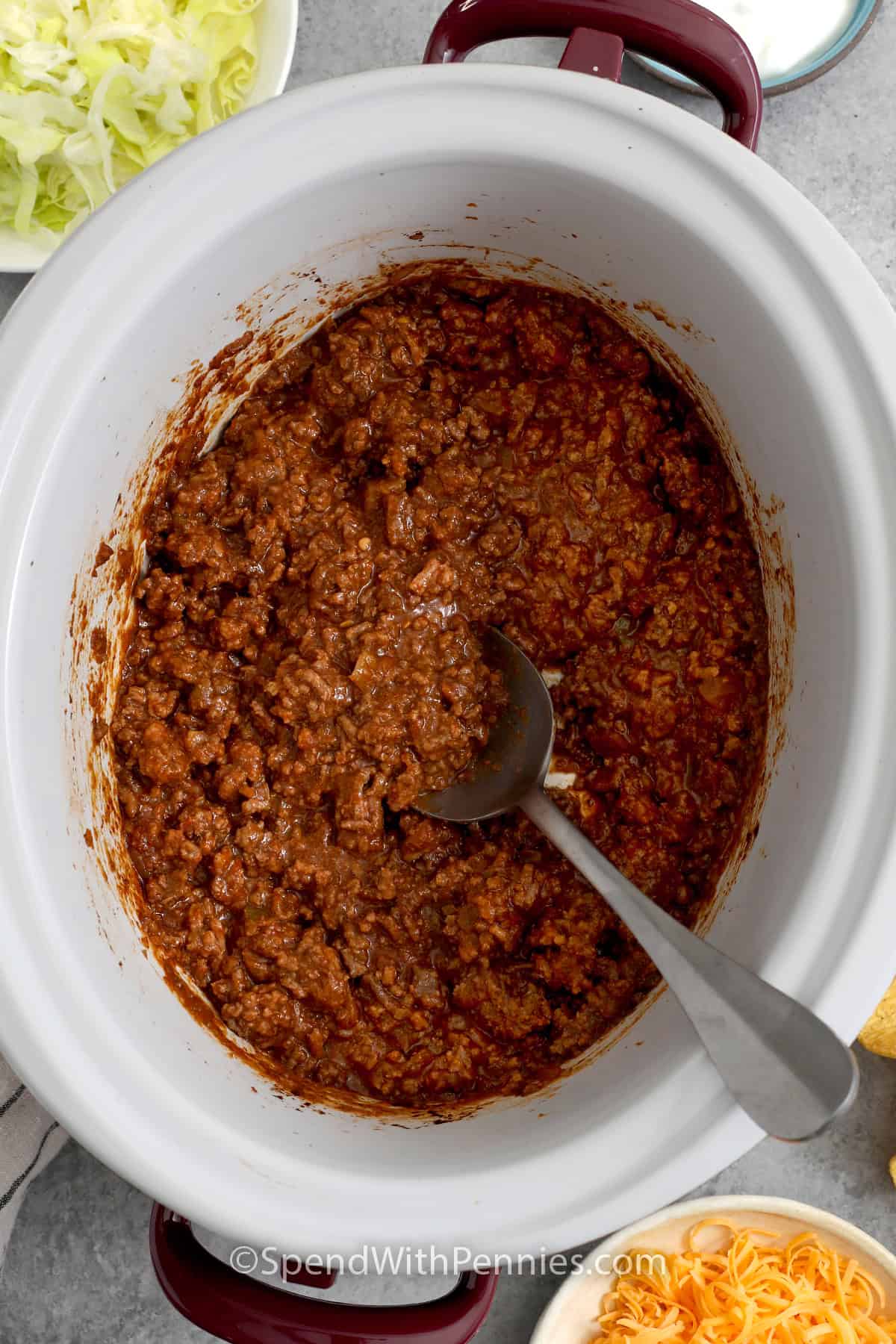 Cooked taco meat in a slow cooker with a spoon and taco toppings off to the side