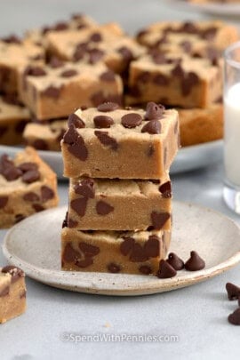 Stack of three Peanut Butter Cookie Bars on a plate with a glass of milk and a tray of bars in the background.