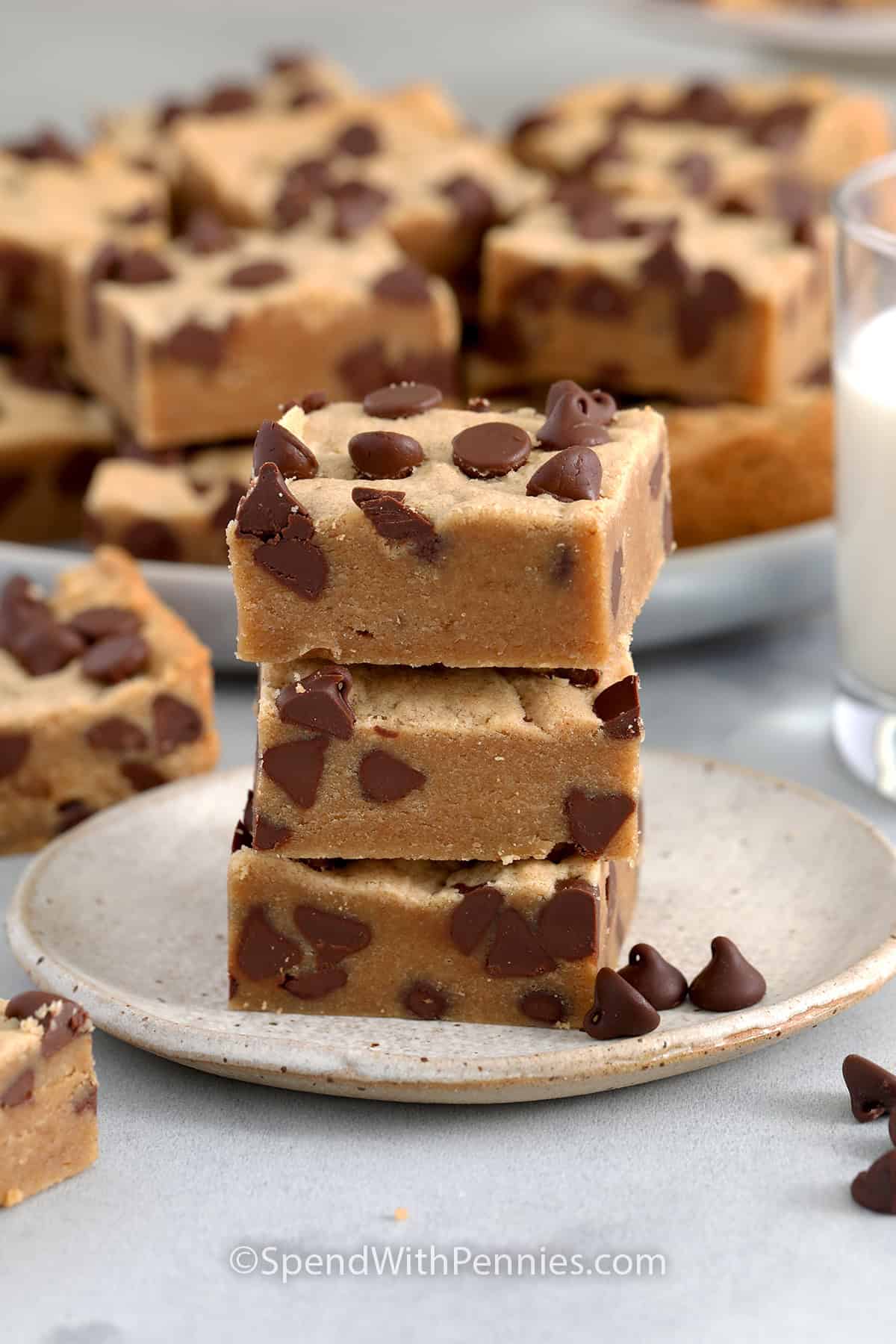 Stack of three Peanut Butter Cookie Bars on a plate with a glass of milk and a tray of bars in the background.