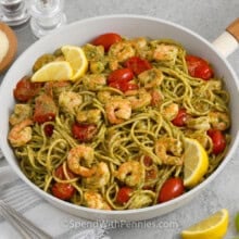 A skillet of Shrimp Pesto Pasta garnished with lemon wedges and parmesan cheese