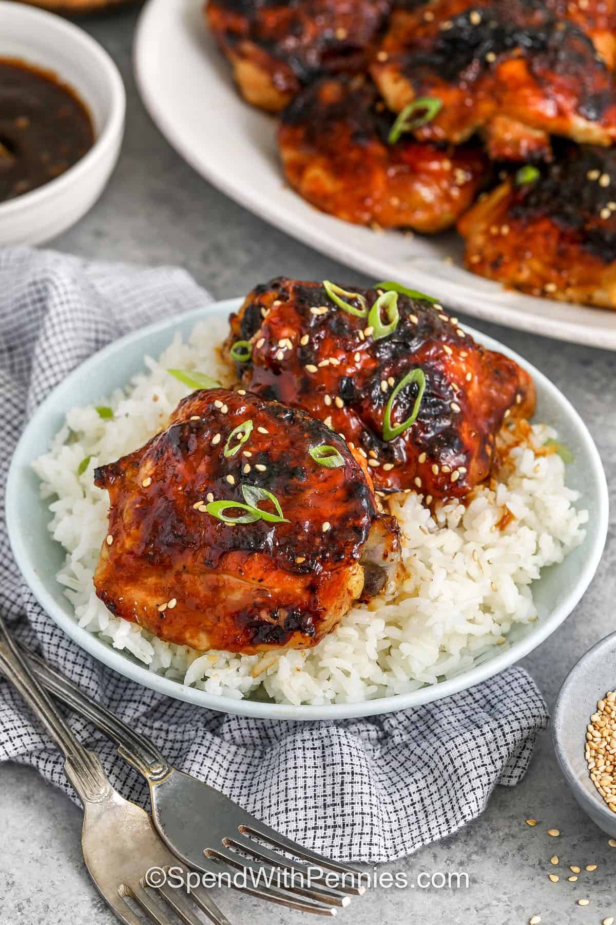These baked chicken thighs are sticky, sweet, and lightly tangy from apple cider vinegar. As they roast, the glaze thickens and coats the skin for full flavor without the extra fuss. With easy ingredients and quick steps, this recipe works for weeknight meals or fun appetizers. Leftovers stay juicy when reheated with a little sauce. #spendwithpennies baked sticky chicken thighs on rice with a platter of chicken thighs in the background