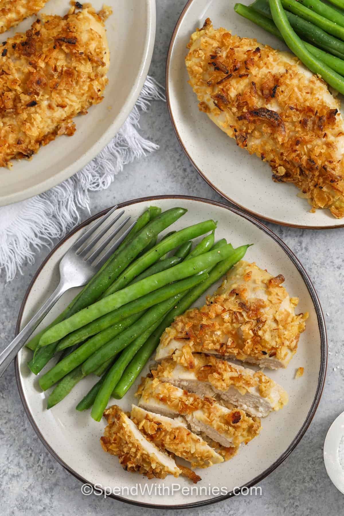 This French onion baked chicken recipe is the easiest way to get a crunchy chicken coating with minimal effort. Crushed crispy fried onions are used as the crust, held in place by an egg and Dijon coating. Baking at the perfect temperature keeps the chicken tender while the topping crisps. For meal prep, cool completely and store up to four days, then reheat in the oven or air fryer for the best crunch. #spendwithpennies french onion baked chicken on a white plate withe beans