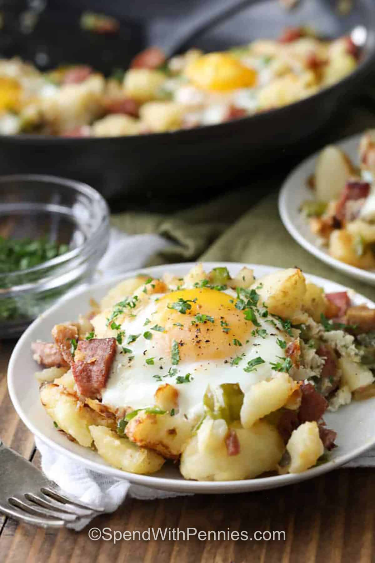 This corned beef hash recipe is the coziest way to use leftover corned beef. Diced russet potatoes are boiled until tender, then skillet-fried in butter with onion and green bell pepper until the edges turn crisp and golden. Crack eggs into wells and finish in the oven for an easy corned beef hash and eggs dinner or brunch. The texture is hearty and satisfying, with salty savory bites in every forkful. #spendwithpennies Corned Beef Hash on a plate and pan full in the back