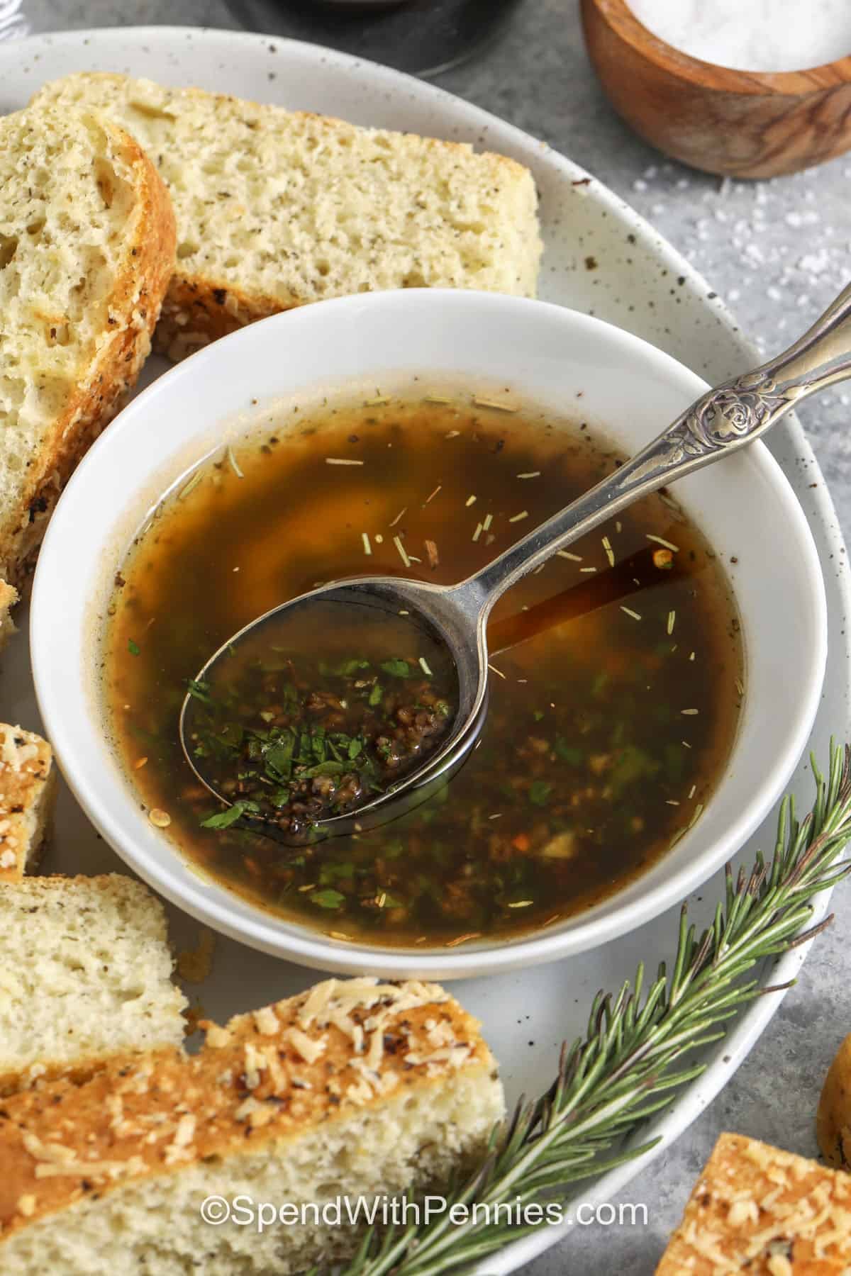 Turn a loaf into an easy appetizer with olive oil bread dip made from pantry seasonings. This bread dipping oil combines extra virgin olive oil with balsamic vinegar, minced garlic, parsley, Italian seasoning, salt, and pepper for a savory, herby finish. Shake, rest, and pour into a shallow dish, then top with Parmesan to serve with warm bread. #spendwithpennies bowl of Olive Oil Bread Dip with a spoon