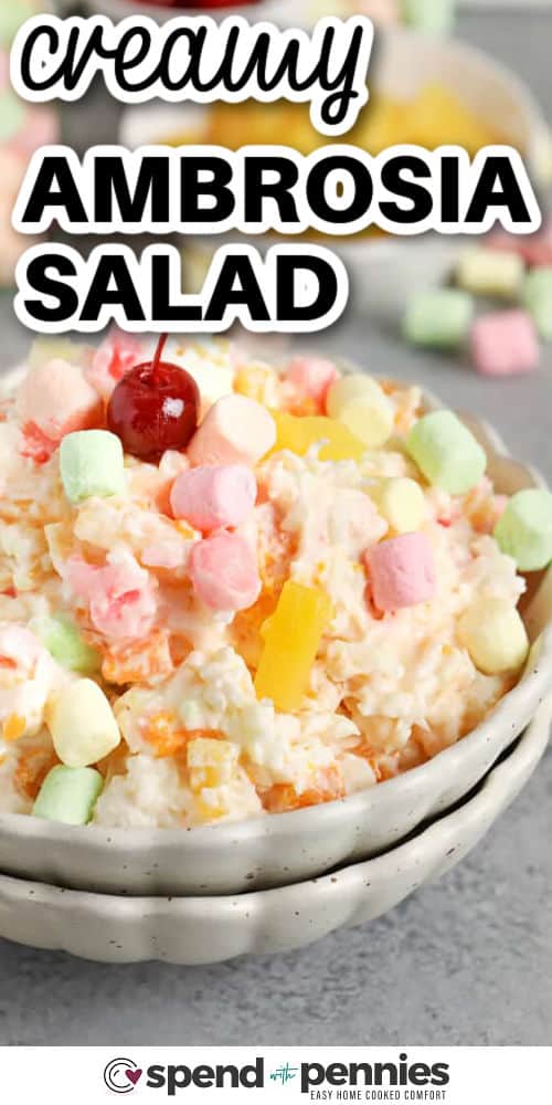 Ambrosia salad is the easiest make-ahead holiday side dish, and it tastes even better after resting in the fridge. The creamy base, coconut, and marshmallows mellow into a fluffy texture while pineapple and mandarin oranges bring bright sweetness. Drain canned fruit well to avoid watery results, and fold oranges in last to keep them intact and pretty for serving. #spendwithpennies creamy Ambrosia Salad in a bowl with writing