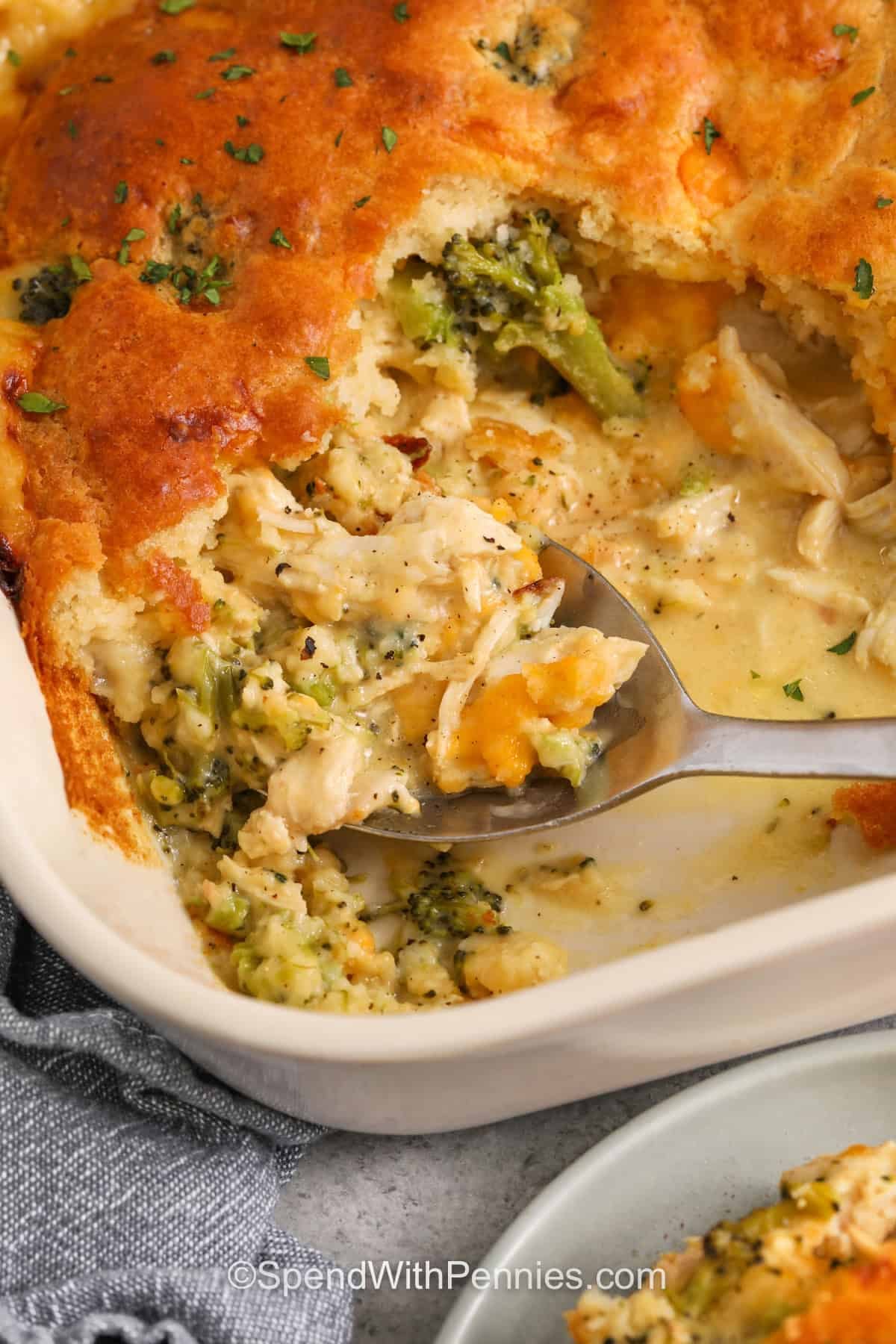 This cheddar chicken cobbler recipe is a hearty baked dinner that balances creamy filling with a fluffy, golden topping. Broccoli gives the dish color and texture, while the cheddar and biscuit mix make it feel a little more special than a standard casserole. It is a family-friendly option for cooler nights, casual gatherings, or any time a one-dish dinner sounds better than cooking separate parts of a meal. #spendwithpennies taking a spoonful of Cheddar Chicken Cobbler out of the casserole dish