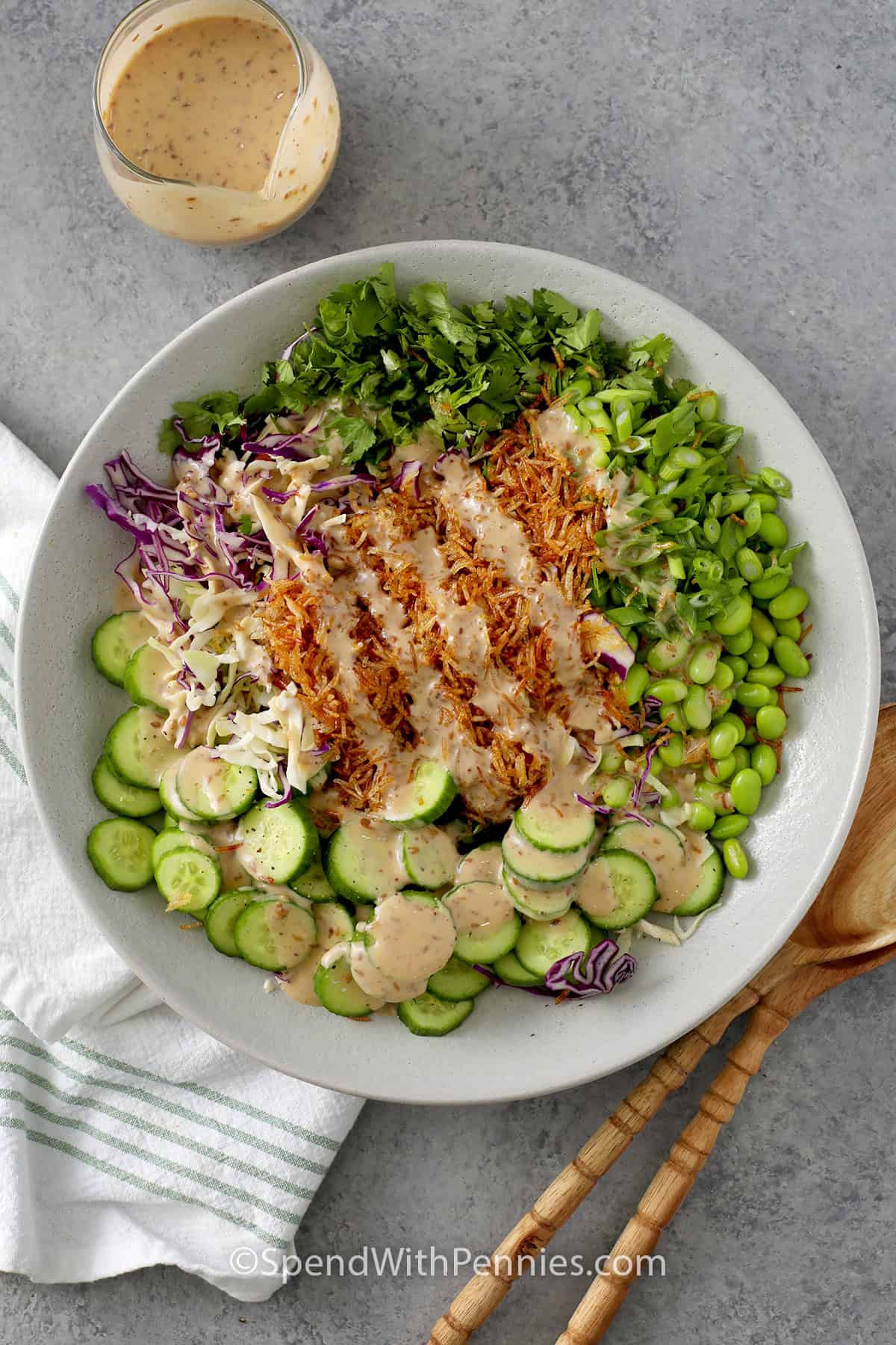A large bowl with chopped cilantro, green onions, edamame, sliced cucumbers, coleslaw mix and crispy rice with dressing poured overtop.
