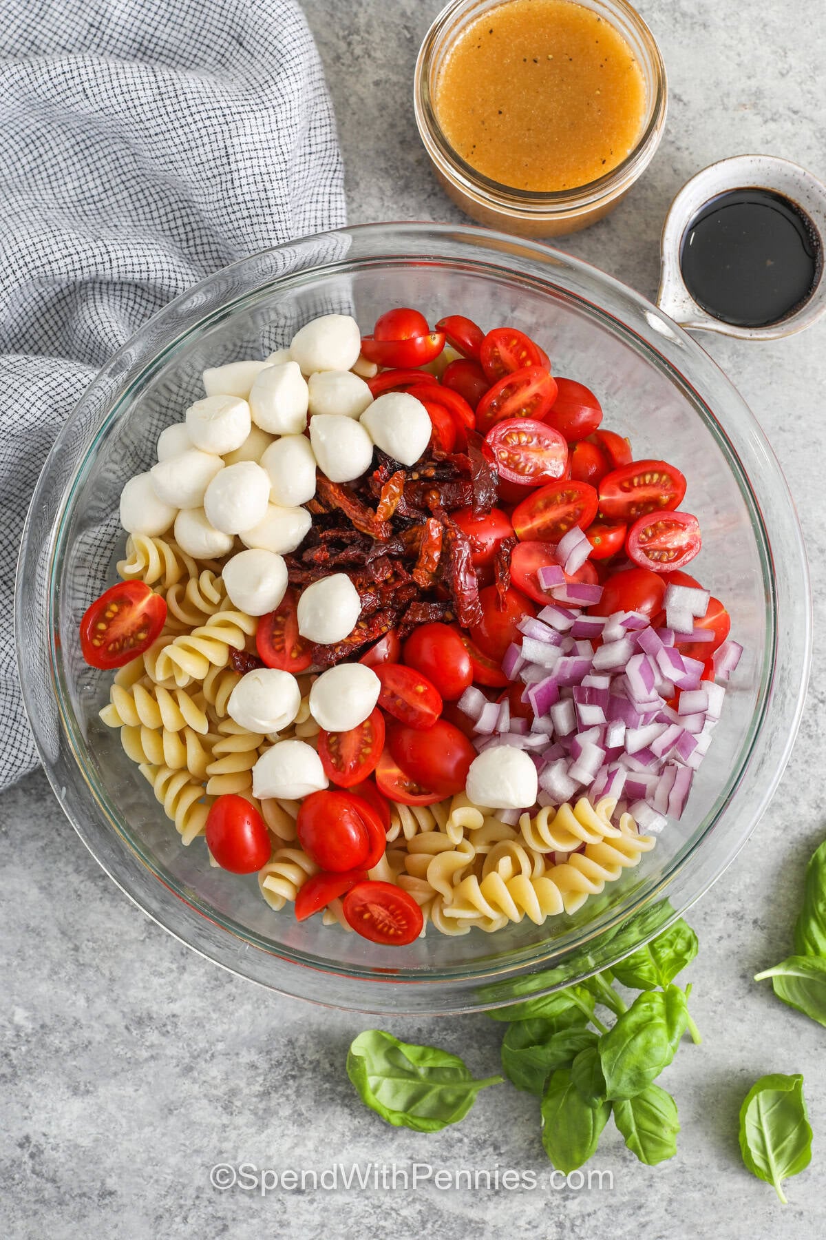 This Caprese pasta salad recipe turns classic tomato, mozzarella, and basil flavors into a hearty chilled pasta salad. Rotini holds the red wine vinaigrette beautifully, while mozzarella pearls and grape tomatoes make each bite creamy and juicy. Make this for warm-weather meal prep because the pasta absorbs flavor as it rests, and the basil stays freshest when added right before serving. #spendwithpennies adding ingredients together in bowl to make Caprese Pasta Salad