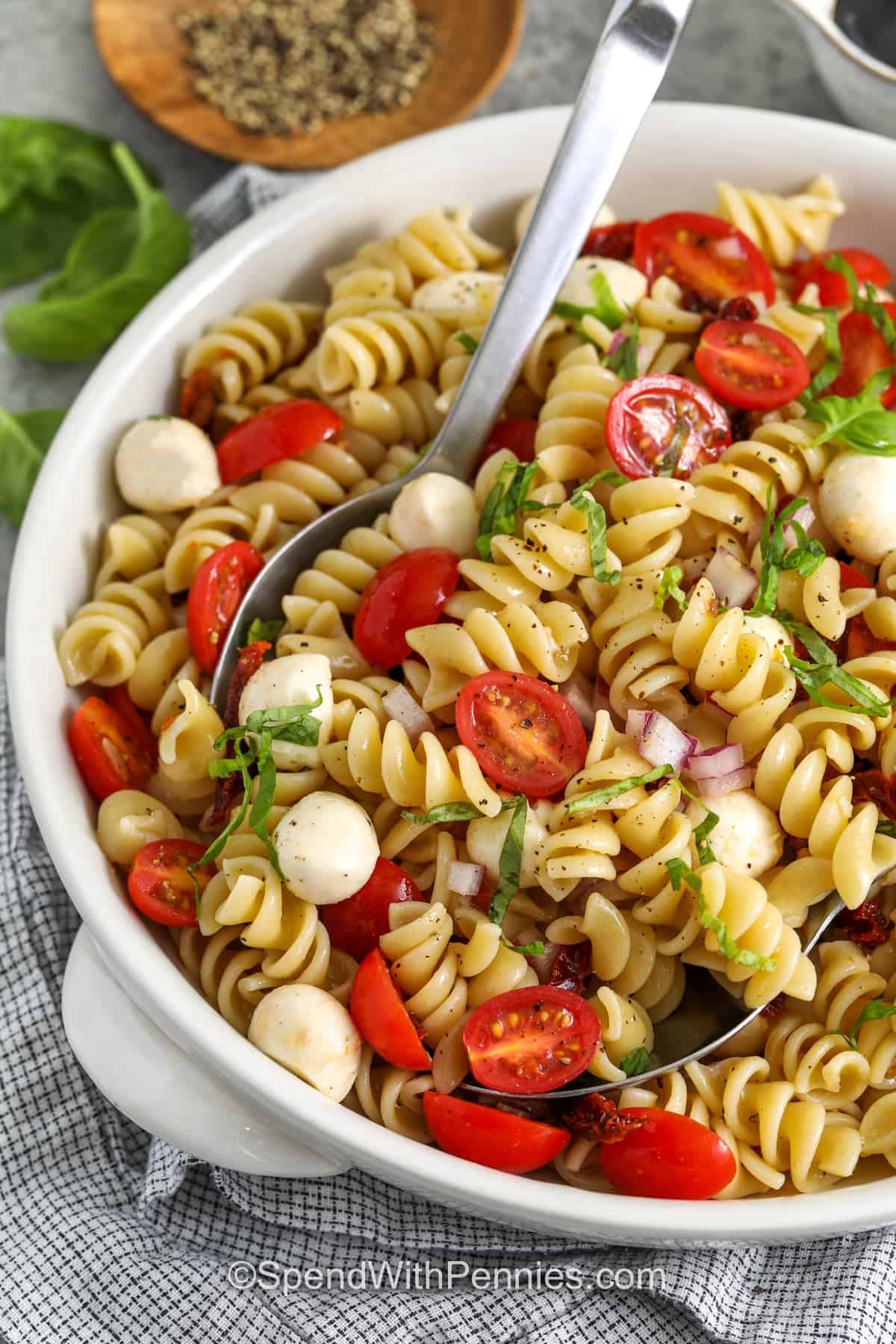 Easy Caprese pasta salad is a bright, chilled side dish made with grape tomatoes, mozzarella pearls, fresh basil, and a tangy red wine dressing. The pasta makes it hearty enough for lunches while still keeping the fresh flavor of a classic Caprese salad. It is a practical recipe for summer parties, backyard BBQs, and simple dinners when you want something cool and flavorful. #spendwithpennies Caprese Pasta Salad in a bowl with spoons