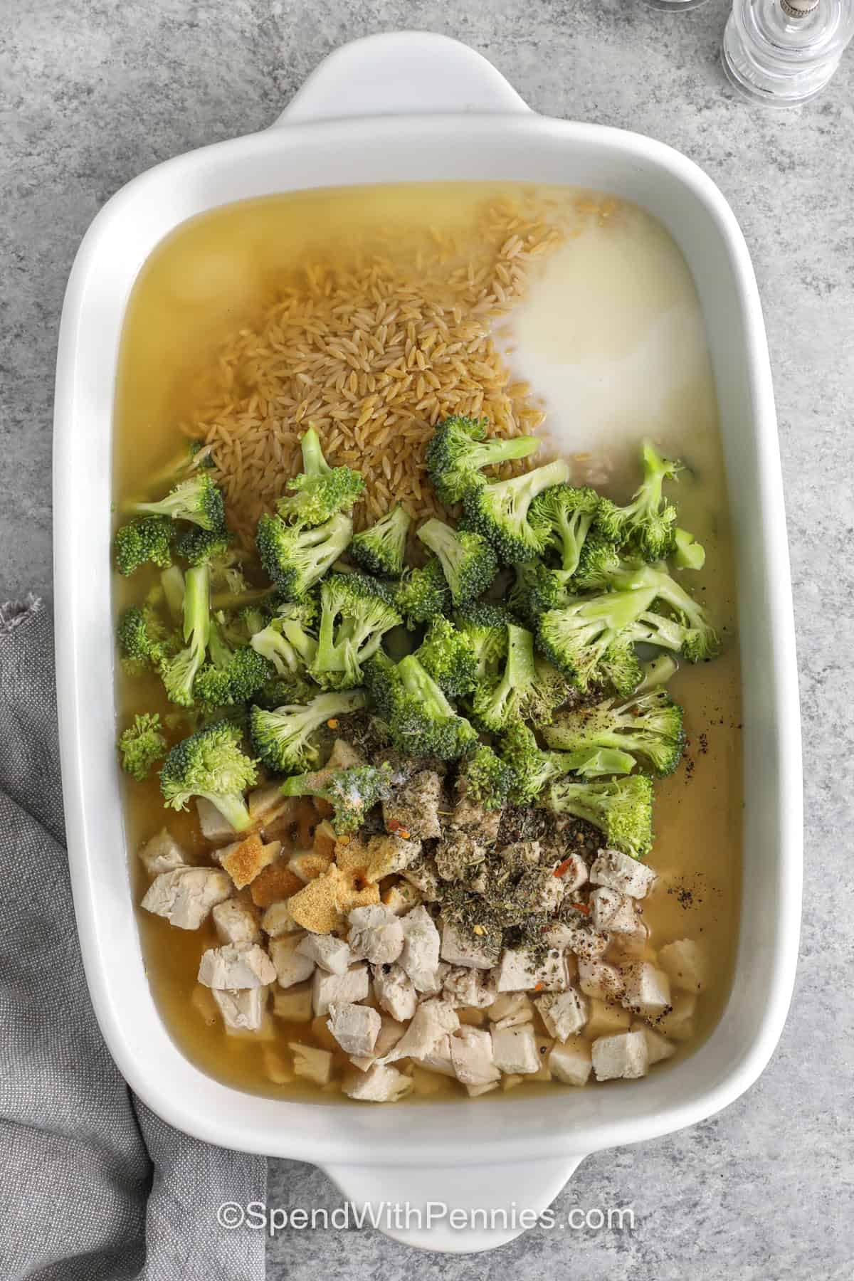 One pan chicken orzo bake is perfect for busy nights when you want a complete meal without a sink full of dishes. The orzo cooks right in the baking dish with chicken, broccoli, broth, and milk, then the cheeses melt into a creamy sauce. Use rotisserie chicken to save time, and stir in a splash of warm broth or milk if the pasta thickens too much after baking. #spendwithpennies adding ingredients to dish to make Chicken Orzo Bake
