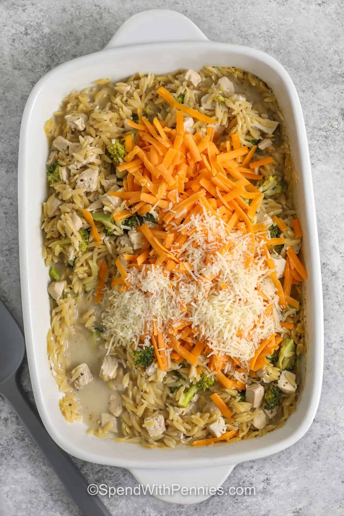 This chicken orzo bake is a comforting one dish dinner made with dry orzo, cooked chicken, broccoli, chicken broth, milk, Parmesan, and cheddar cheese. The orzo bakes right in the pan, soaking up the creamy liquid as it cooks, so there is no need to boil it first. It is a practical weeknight casserole with tender pasta, melty cheese, and a veggie tucked into every scoop. #spendwithpennies adding cheese to casserole dish to make Chicken Orzo Bake