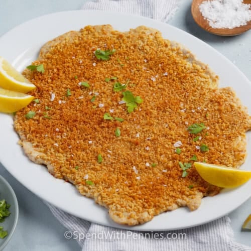 Horizontal view of a giant chicken cutlet on a serving plate with lemon.