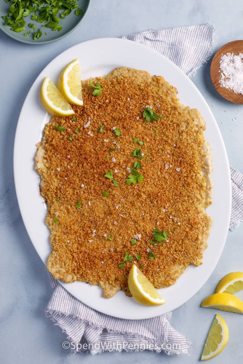 Overhead of giant chicken cutlet on a serving plate with lemon and flakey salt.