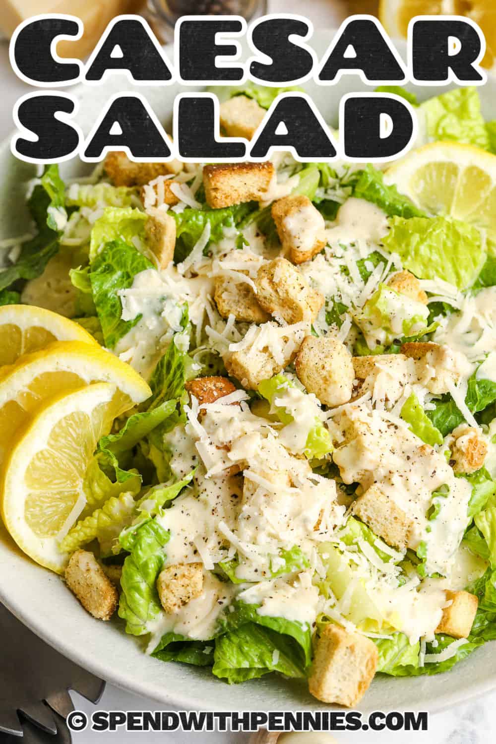 This Caesar salad is crisp, creamy, and full of classic flavor from fresh romaine, crunchy croutons, Parmesan cheese, and a homemade Caesar dressing. The blender dressing is made with garlic, lemon juice, Dijon, Worcestershire, anchovy, capers, egg, and mild oil for a rich texture that coats every leaf. Serve it with pasta, steak, chicken, or seafood for an easy side dish that feels special. #spendwithpennies Caesar Salad with dressing and croutons on top with a title