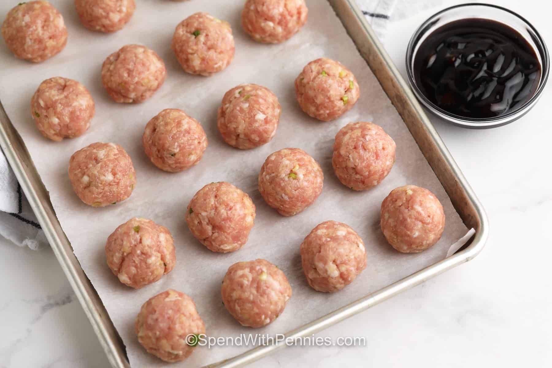 Hoisin meatballs with ginger and green onions are ready in just over 30 minutes and can be served so many ways. Bake the pork meatballs until tender, simmer the sweet and savory hoisin sauce, and toss everything together for a cozy, saucy recipe that is freezer-friendly and great for leftovers. #spendwithpennies top view of uncooked meatballs on a parchment lined pan with a bowl of hoisin glaze