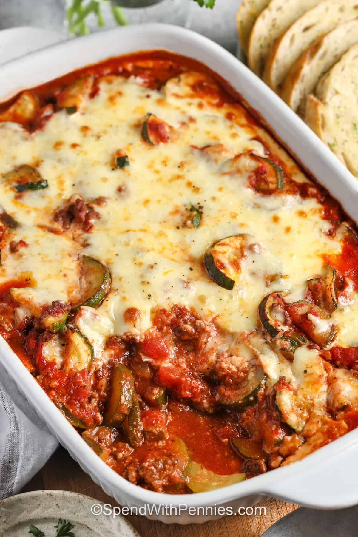 This zucchini hamburger casserole stretches one pound of beef with several fresh zucchini, tomatoes, and flavorful pasta sauce. The casserole is hearty enough to serve alone, yet it can also be spooned over pasta, rice, or quinoa when feeding a larger group. Its saucy center and browned cheese topping make leftovers worth looking forward to. Save this simple dinner for busy days and meal prep. #spendwithpennies cheesy Zucchini Hamburger Casserole in the casserole dish with a portion taken out