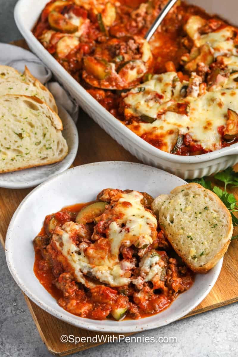 Easy zucchini hamburger casserole combines fresh vegetables with the cozy flavors of a cheesy tomato and beef bake. A simple egg and milk mixture coats the Italian cheese, helping the topping bake into a golden layer that stays with every serving. Cook the zucchini briefly for a firmer texture or longer for a softer bite. Save this flexible family casserole for a relaxed weeknight dinner. #spendwithpennies Zucchini Hamburger Casserole in the casserole dish and plated with garlic toast