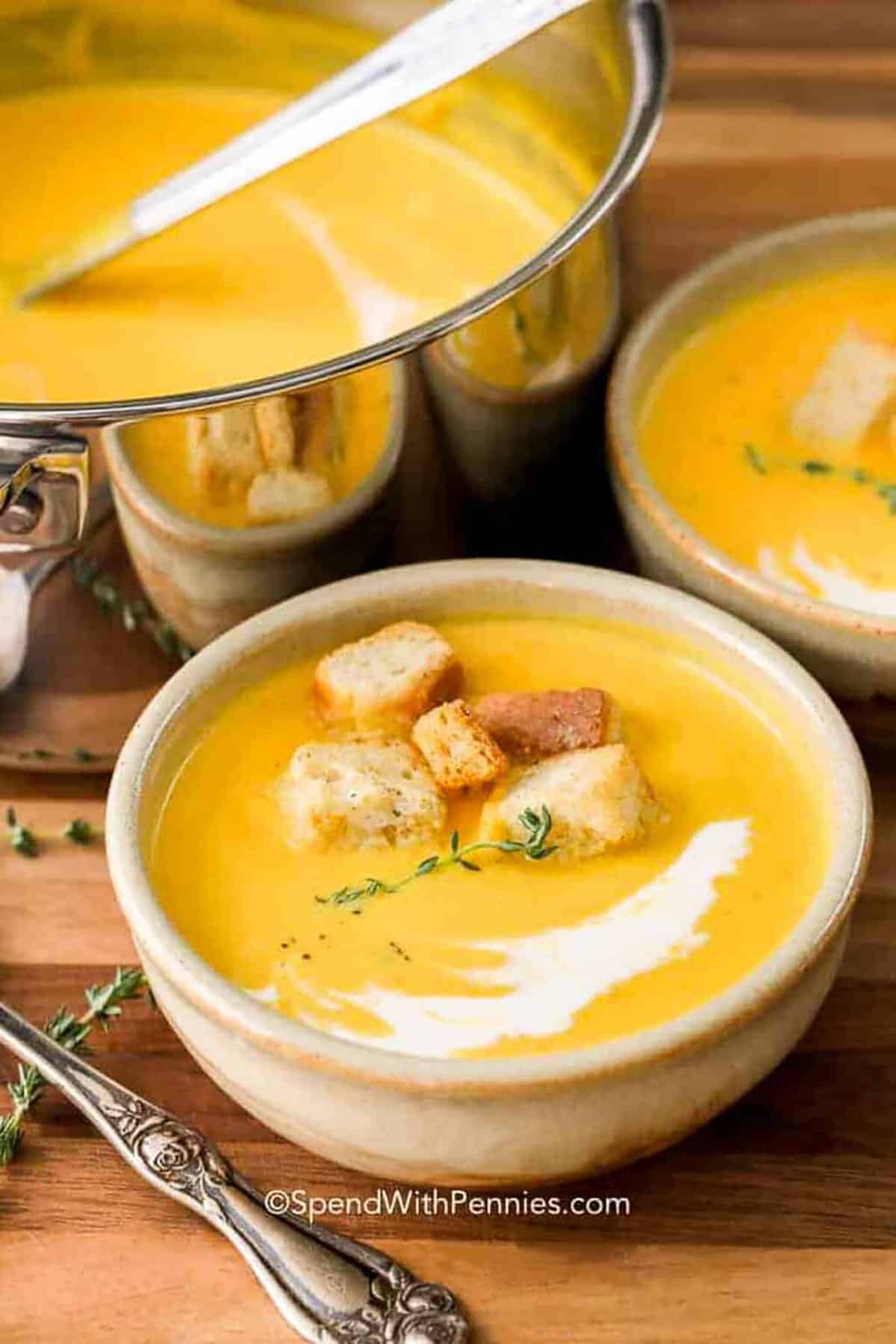 Roasted butternut squash soup is an easy way to turn a simple winter squash into a rich and comforting meal. The squash is roasted until tender, then blended with apple, chicken broth, sage, thyme, and cream for a velvety finish. Roasting in large pieces also saves the work of peeling and cubing hard raw squash. Save this soup for your next cozy fall or winter dinner. #spendwithpennies bowls of Butternut Squash Soup with pot full in the back