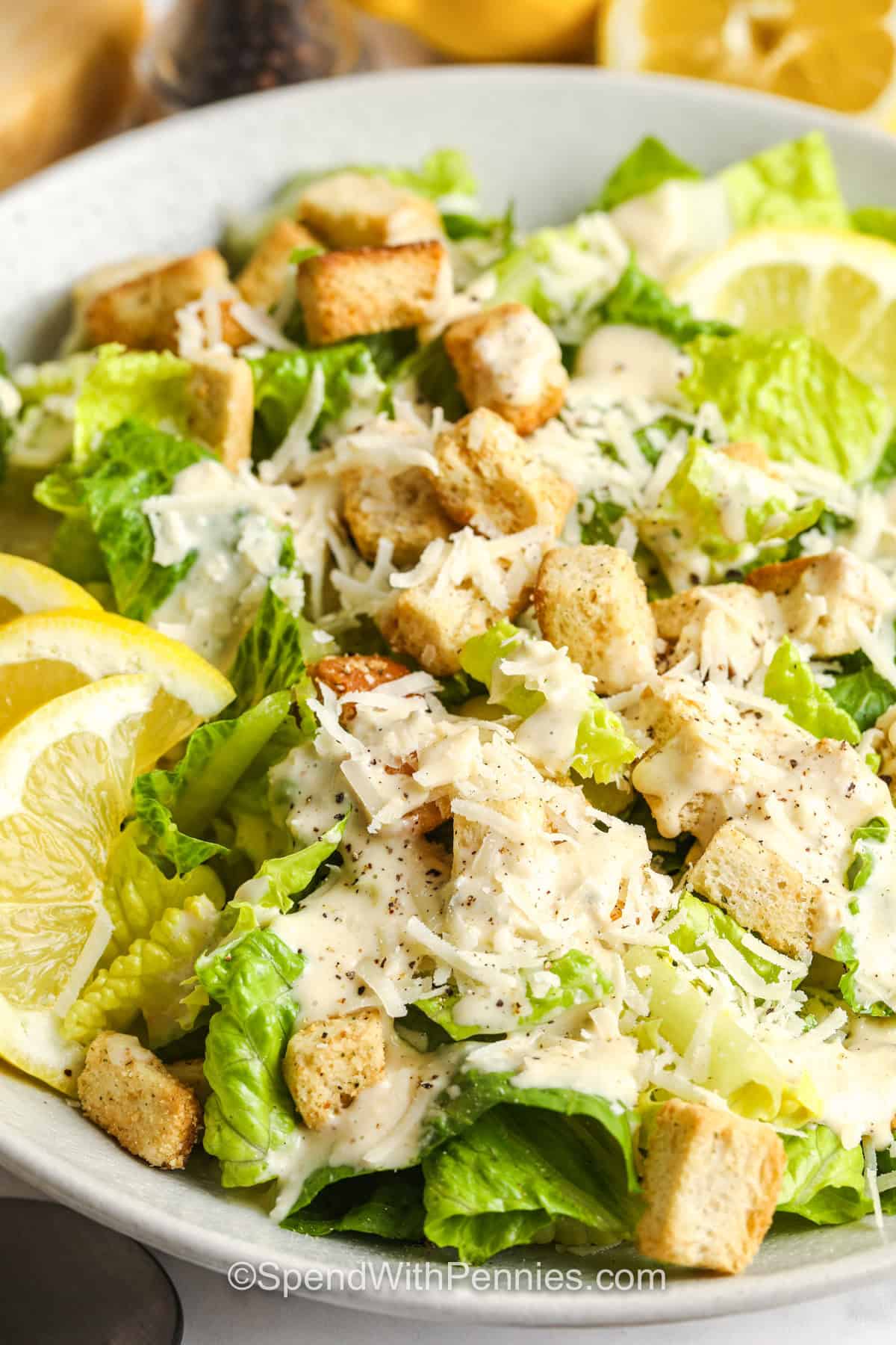 Make classic Caesar salad at home with crunchy romaine lettuce, crisp croutons, Parmesan cheese, and a thick homemade Caesar dressing. Slowly blending light olive oil with egg, lemon, Dijon, garlic, and anchovies creates a smooth, creamy texture without mayonnaise. It's a simple 25-minute side for steak, chicken, pasta, or a holiday dinner. Save this classic salad recipe when you need a dependable side dish. #spendwithpennies Caesar Salad with lemon slices and croutons .