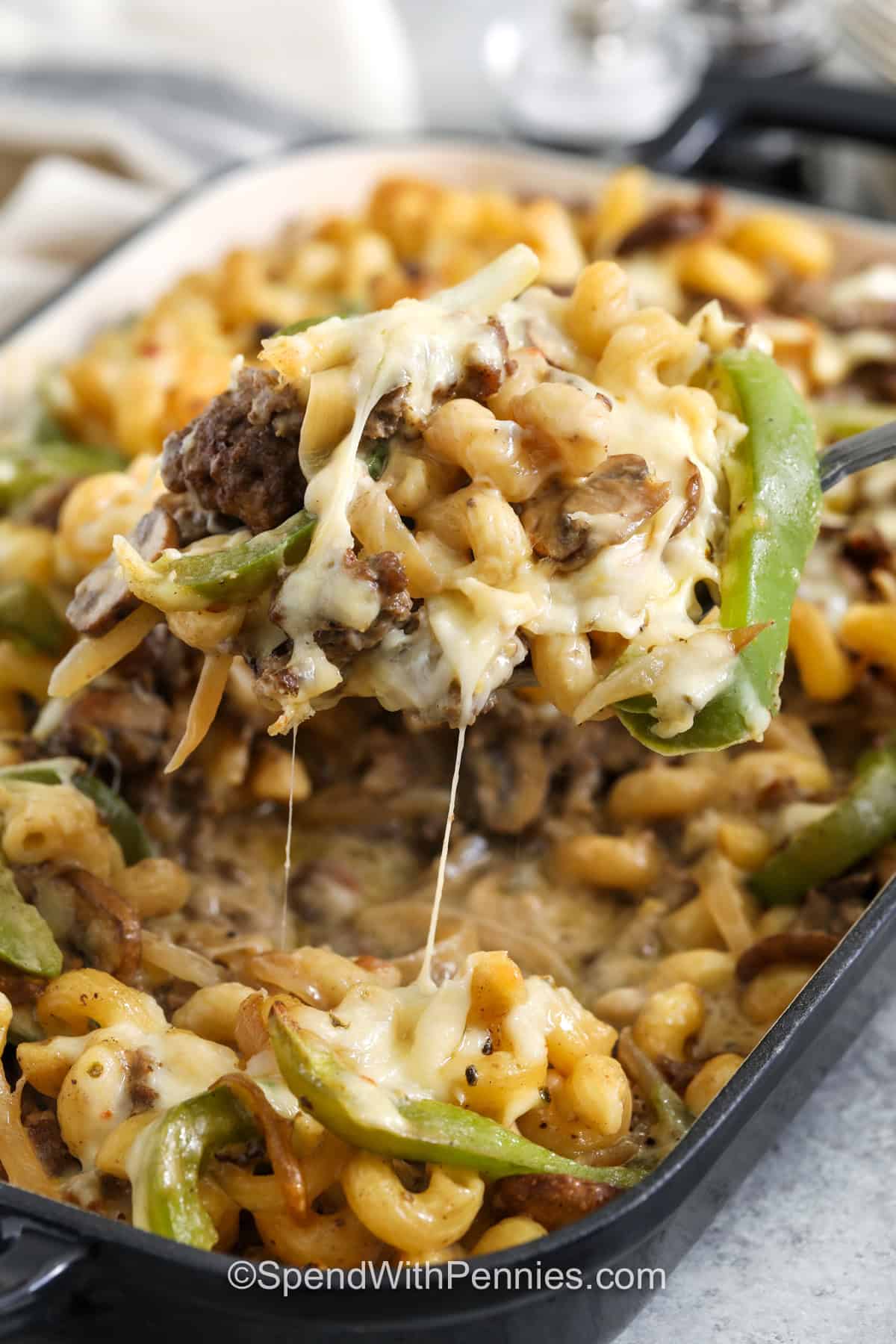 This Philly cheesesteak casserole is an easy way to make six hearty servings with one pound of ground beef. Seasoned beef, green pepper, mushrooms, and onions are mixed with cavatappi and a smooth homemade cheese sauce, then topped with pepper jack and baked until bubbly. The combination is creamy, savory, and satisfying without requiring steak. Pin this practical pasta casserole for family dinners and meal planning. #spendwithpennies taking a spoonful of Philly Cheesesteak Casserole out of the dish.
