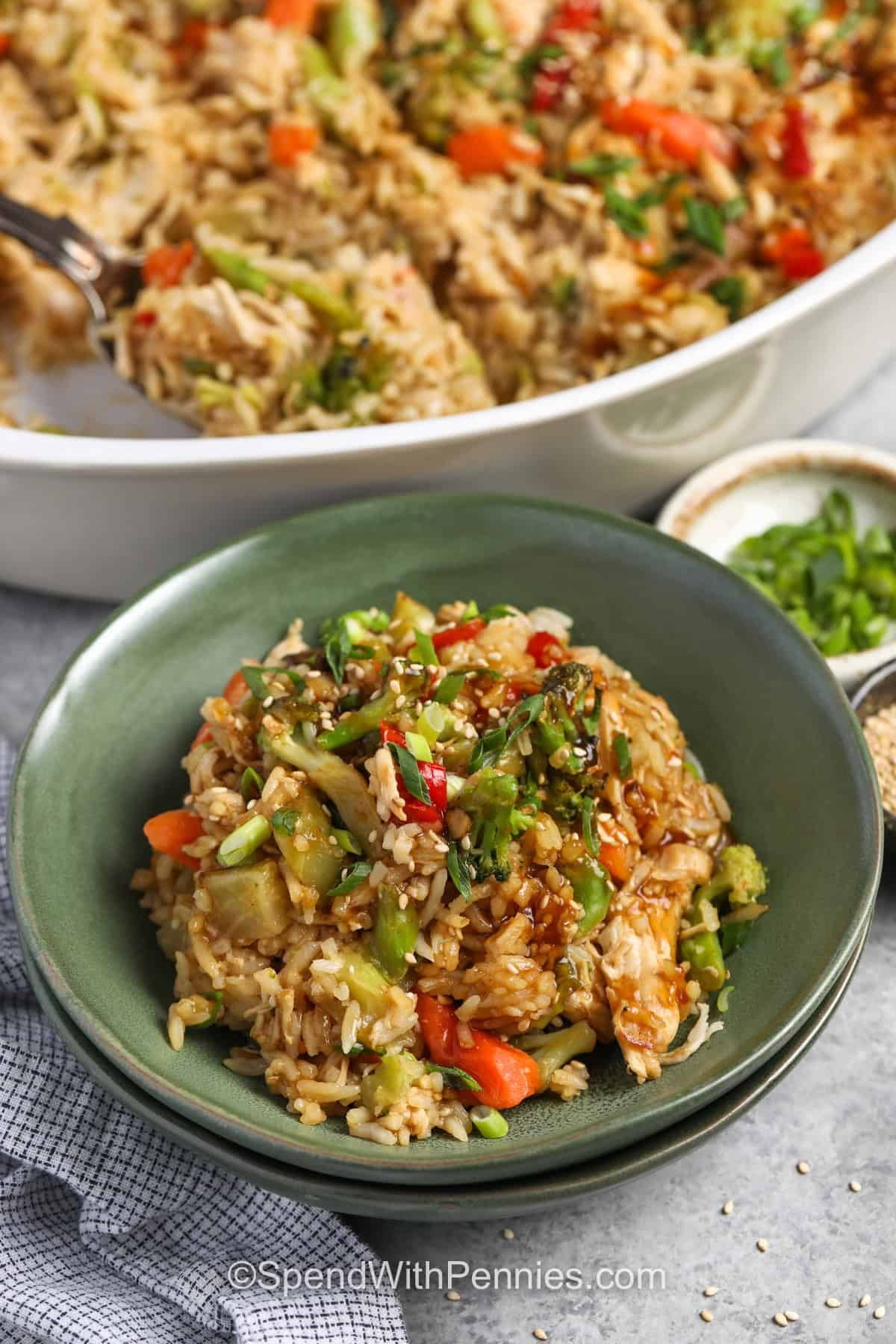 Easy teriyaki chicken casserole makes a warm, satisfying dinner without spending the evening chopping vegetables. Cooked chicken and rice are combined with frozen stir-fry vegetables and a sweet, savory ginger teriyaki sauce, then baked until everything is hot and flavorful. A little reserved sauce drizzled over the casserole at the end keeps every serving saucy. Save this simple recipe for nights when dinner needs to be easy. #spendwithpennies plated Teriyaki Chicken Casserole with pan full in the back.