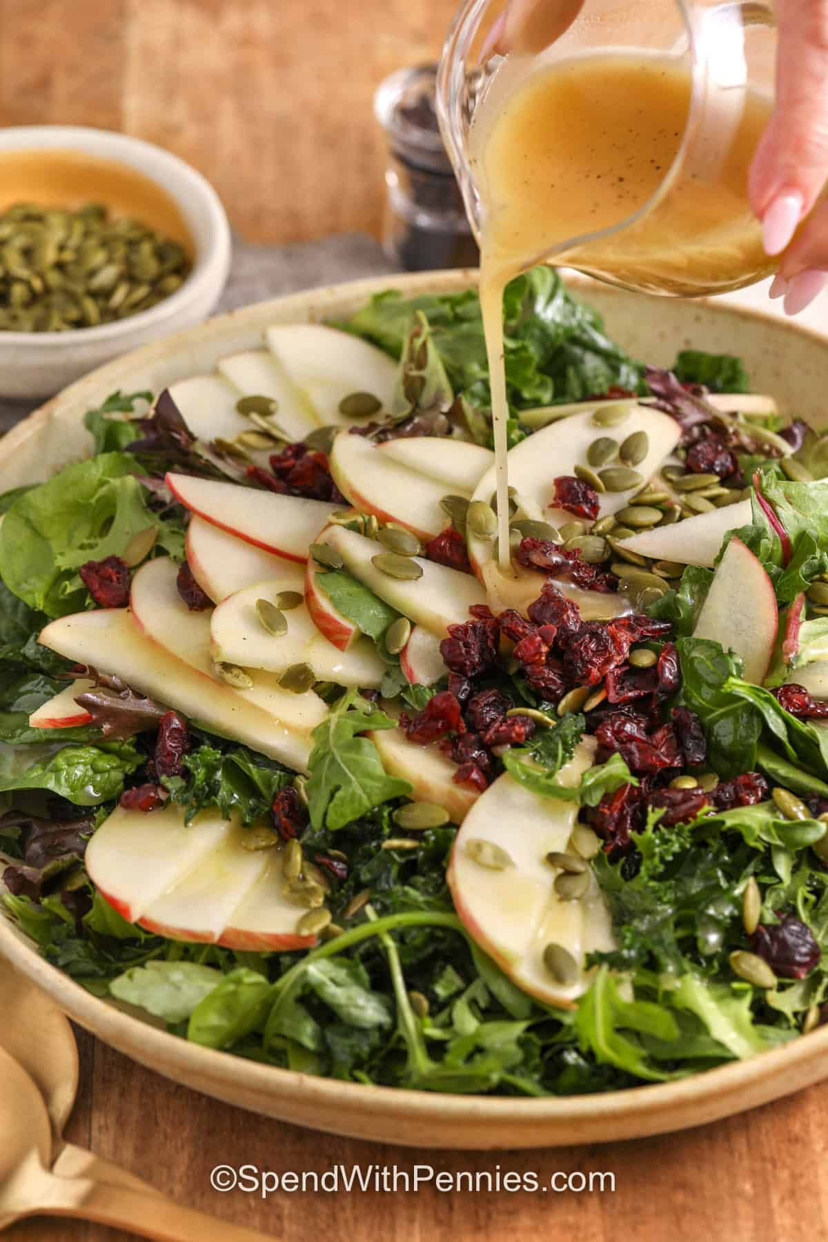 An easy Thanksgiving salad is the perfect fresh contrast to turkey, stuffing, gravy, and creamy potatoes. This version combines thinly sliced apples, kale, arugula, dried cranberries, and crunchy pumpkin seeds with a honey-Dijon apple cider vinaigrette. Massaging the kale makes it tender, while a simple salt-water soak helps the apple slices stay fresh-looking. Add this colorful fall side to your holiday menu and save the recipe. #spendwithpennies Thanksgiving salad in a bowl with a far of dressing.